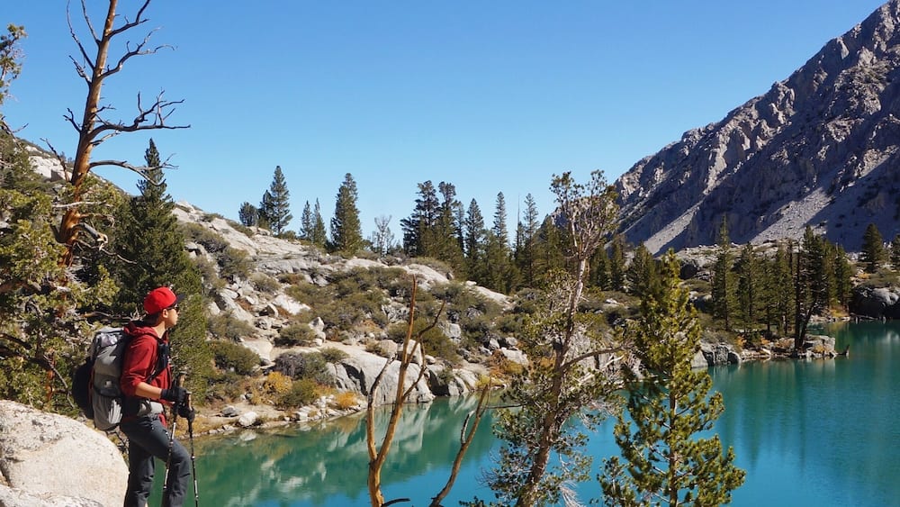 A hiker on a rocky outcrop overlooking a turquoise lake surrounded by trees and mountains.