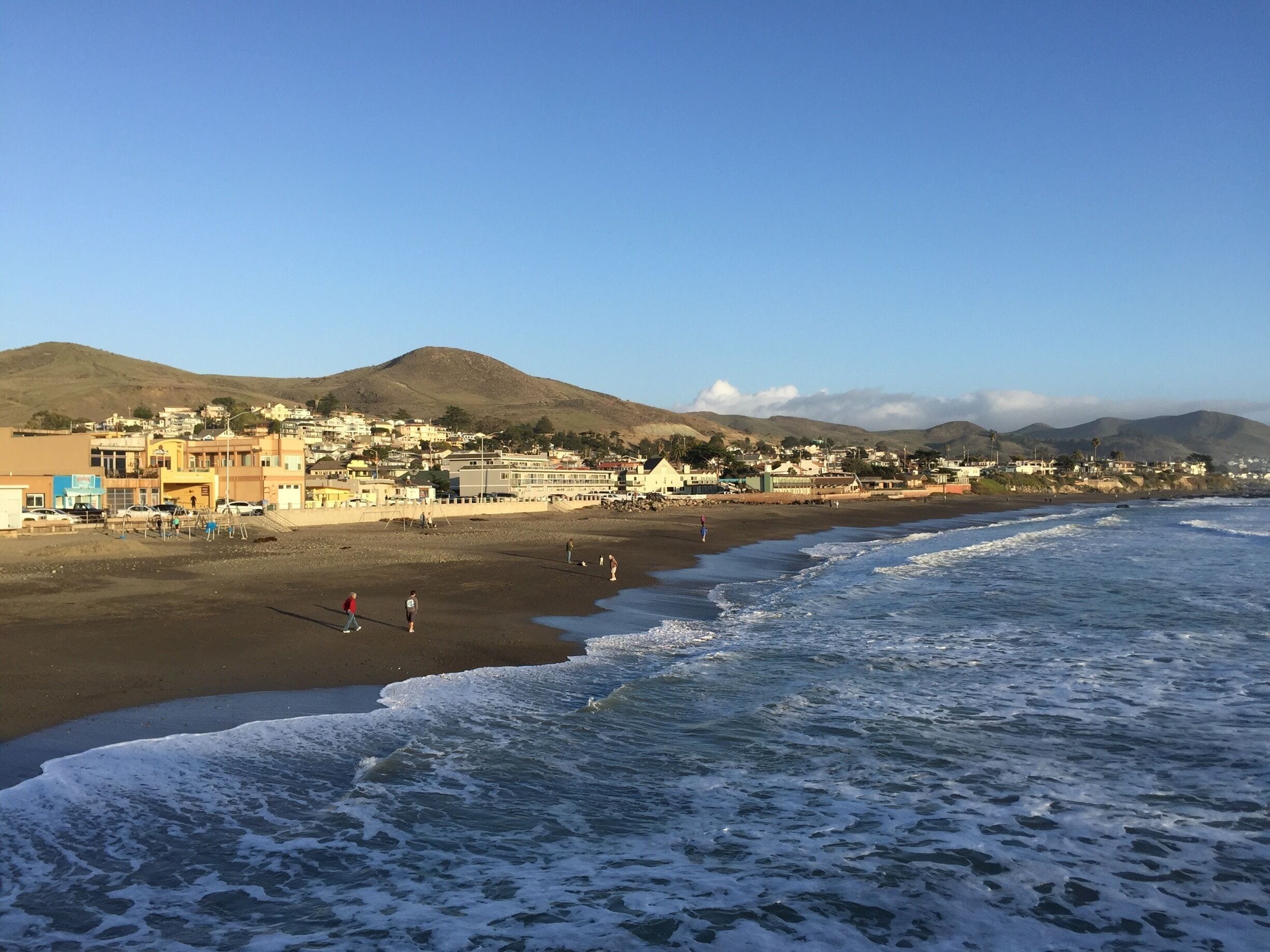 A coastal town with a beach, ocean waves, and hills in the background.