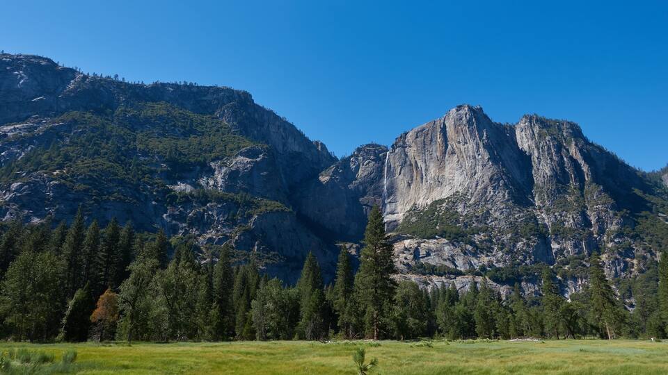 A mountain landscape with a prominent rocky peak, a forested area in the foreground, and a clear blue sky.