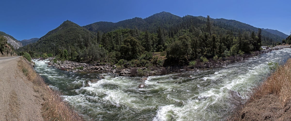 A fast-flowing river with rocky banks, surrounded by dense forest and mountains.