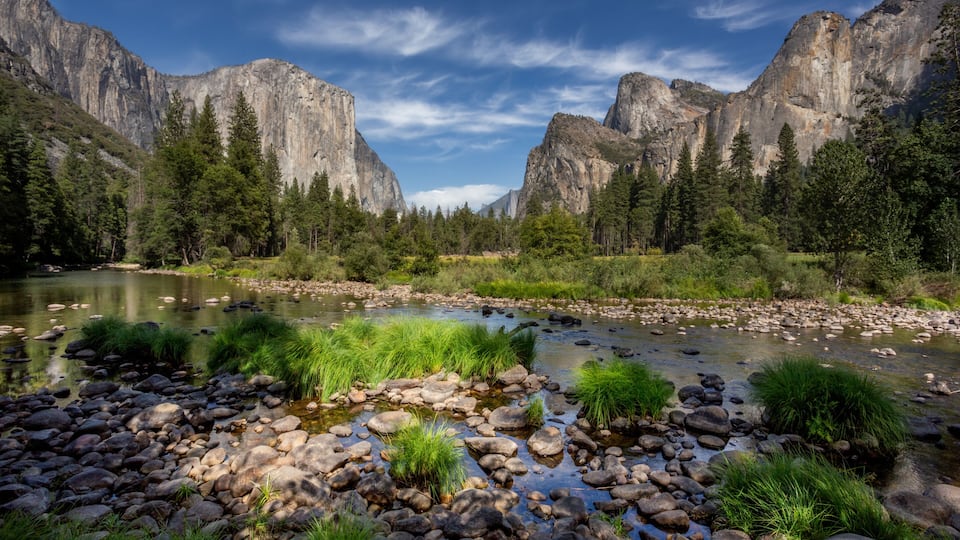 A river with rocks and grass, surrounded by mountains and trees.