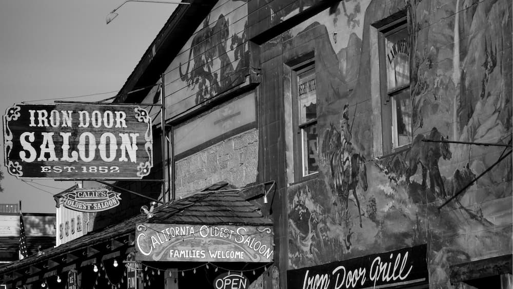 A black and white photo of a historic building with signs for 'Iron Door Saloon' and 'California Oldest Saloon'.