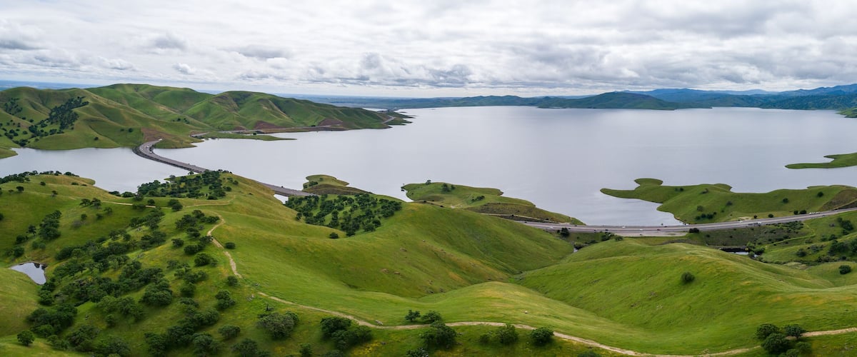A lake surrounded by green hills, a winding road, and distant mountains.