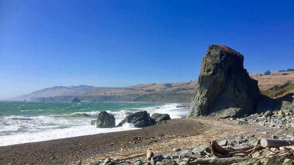 A rocky beach with waves crashing against the shore, a prominent rock formation, and distant hills under a clear blue sky.