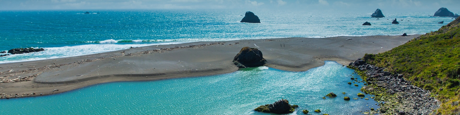 Paisagem costeira com águas azul-turquesa, uma praia de areia e formações rochosas.