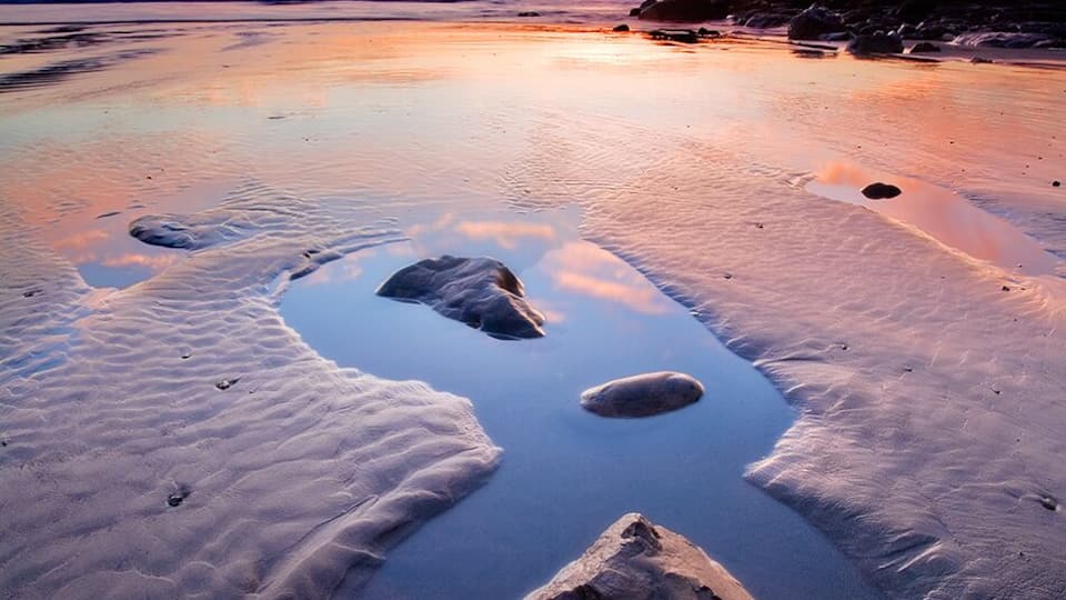A beach with three large rocks in the sand and a sunset in the background.