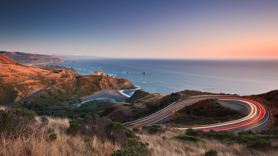 A winding coastal road with a sunset view of the ocean and cliffs.