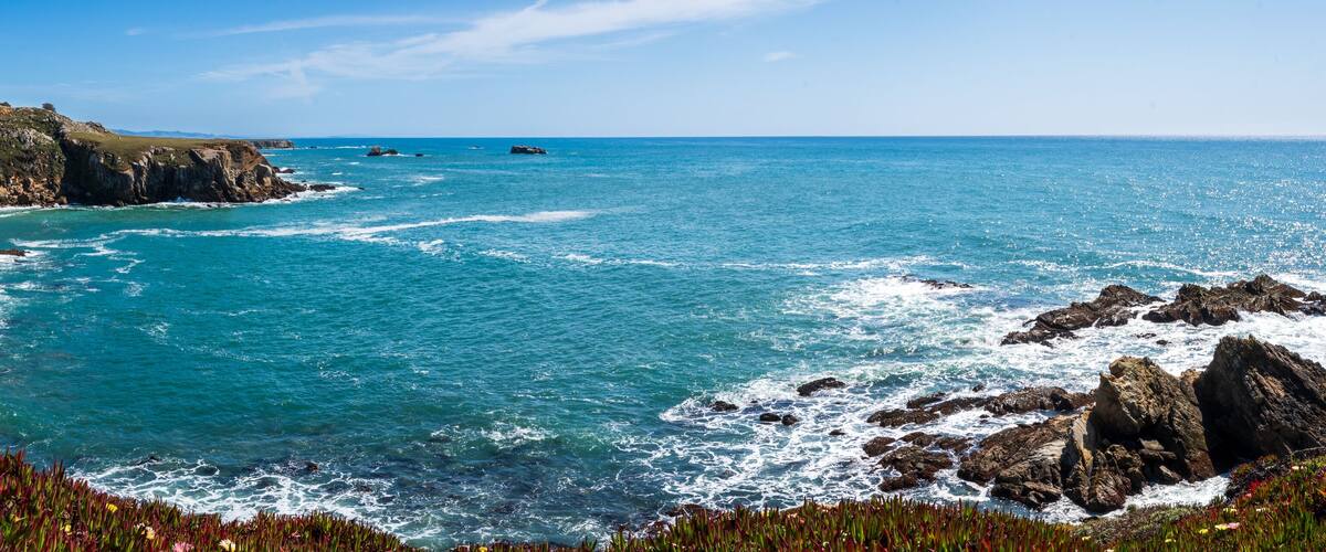 A coastal landscape with rocky cliffs, a sandy beach, and a clear blue sea.