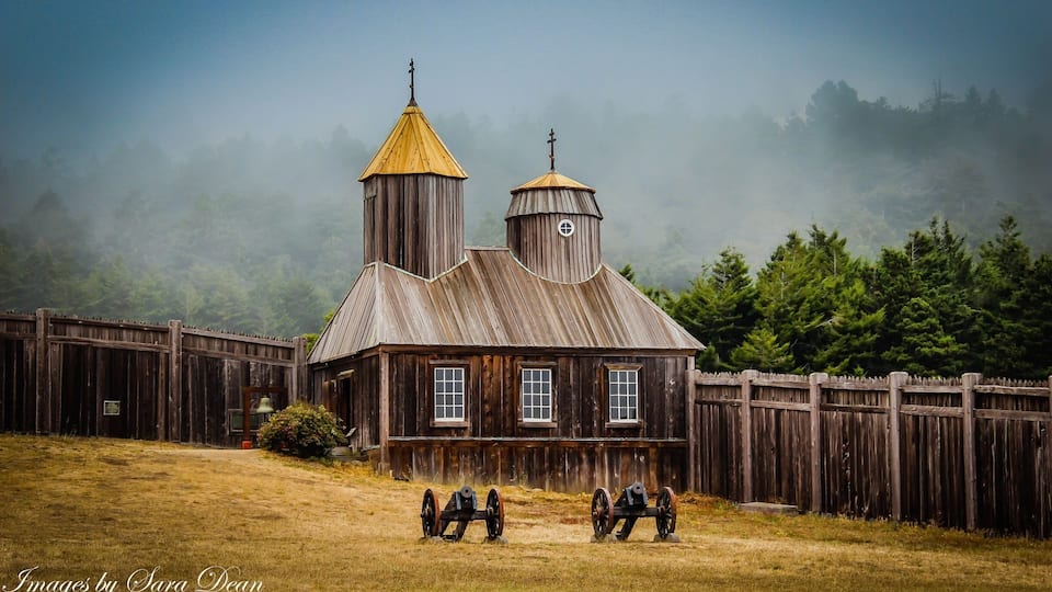 A rustic wooden building with a bell tower and a fence in a foggy forest.