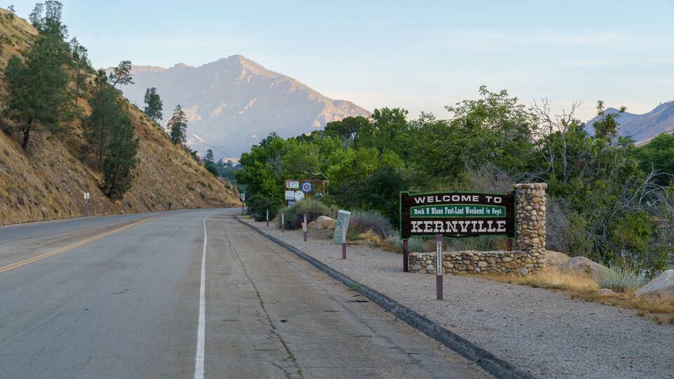 A road with a welcome sign to Kehrville, surrounded by mountains and dry vegetation.