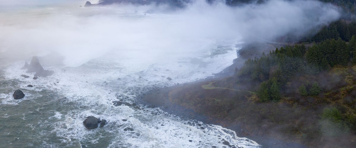A coastal landscape with waves crashing against rocks and a forested cliff.