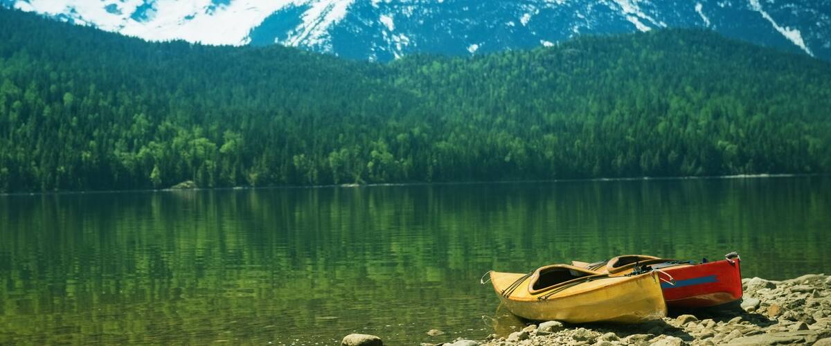 A calm lake with two boats on the shore, surrounded by dense forest and mountains in the background.