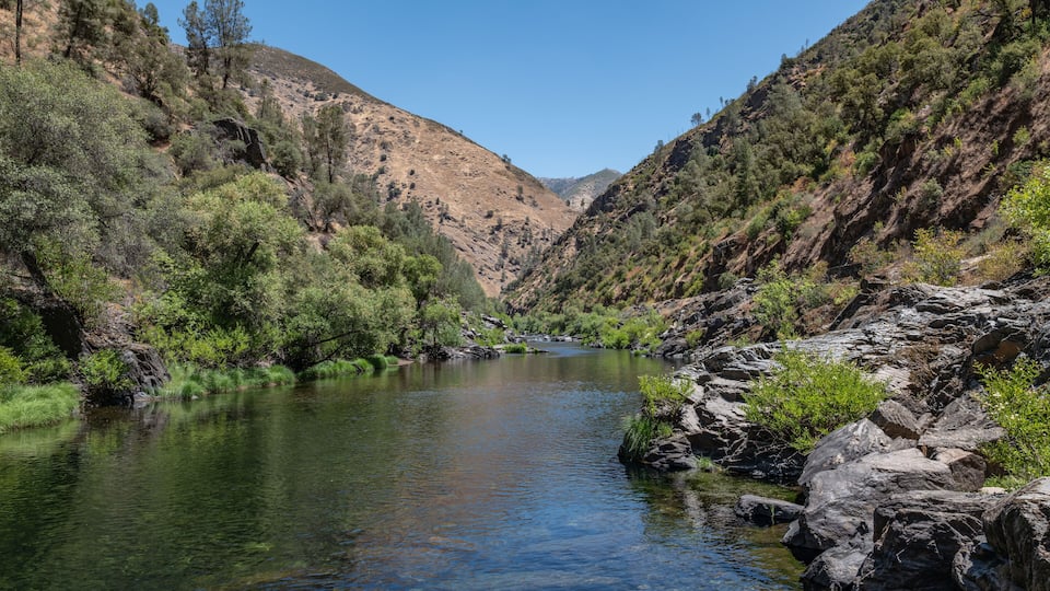 A clear river winding through a rocky canyon with sparse vegetation and hills in the background.