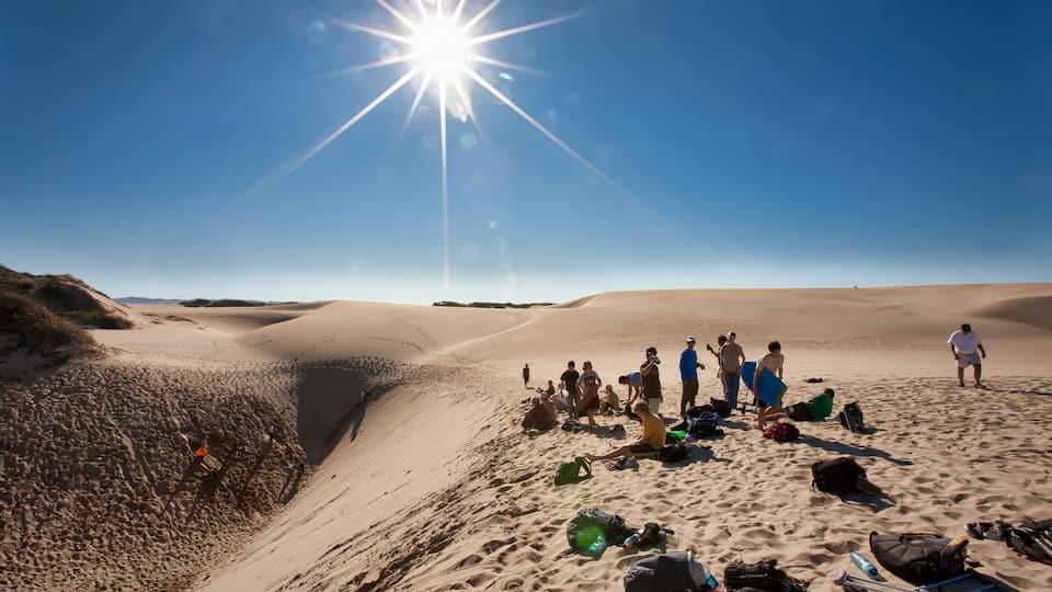 A desert landscape with sand dunes, a group of people with backpacks, and a bright sun in the sky.