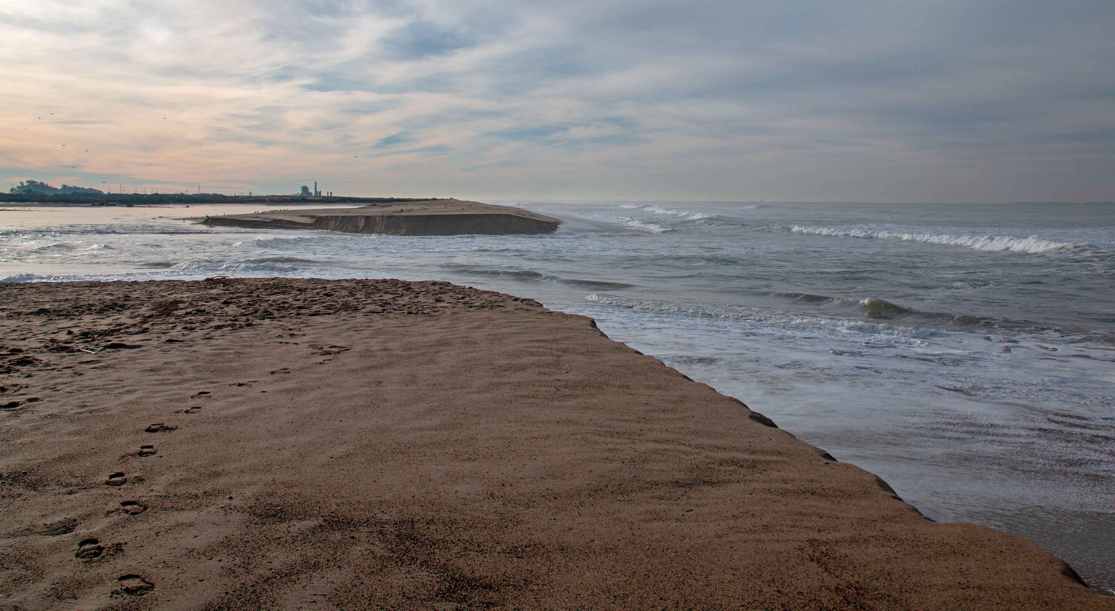 A beach with footprints leading to the water, waves crashing onto the shore, and a distant city skyline.