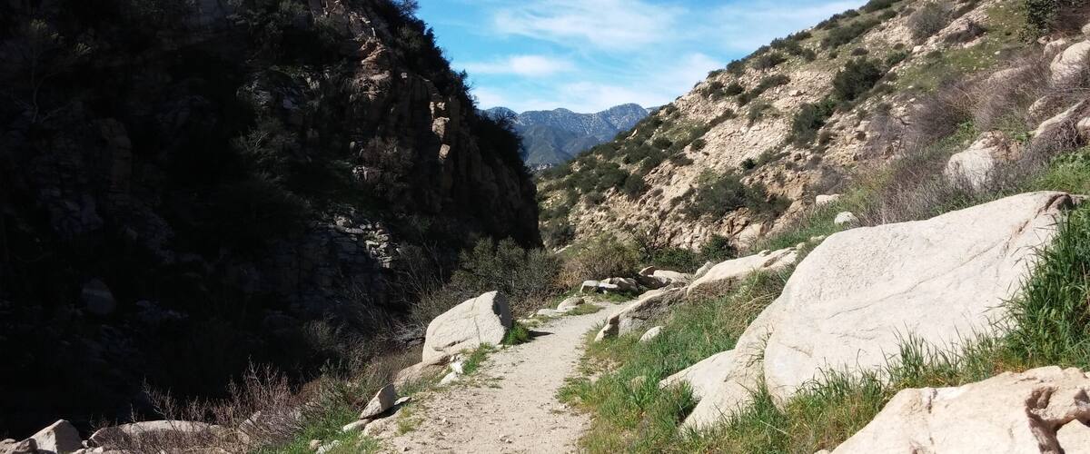 A narrow dirt path flanked by large boulders and rocky cliffs, leading towards distant mountains under a clear sky.