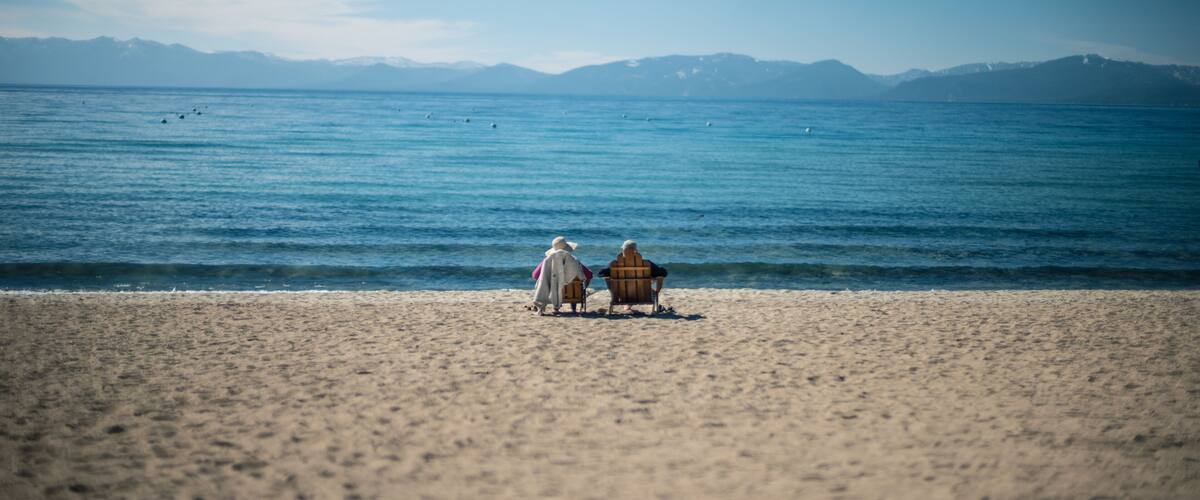 A beach with two people sitting on chairs, a wooden hut, and mountains in the background.