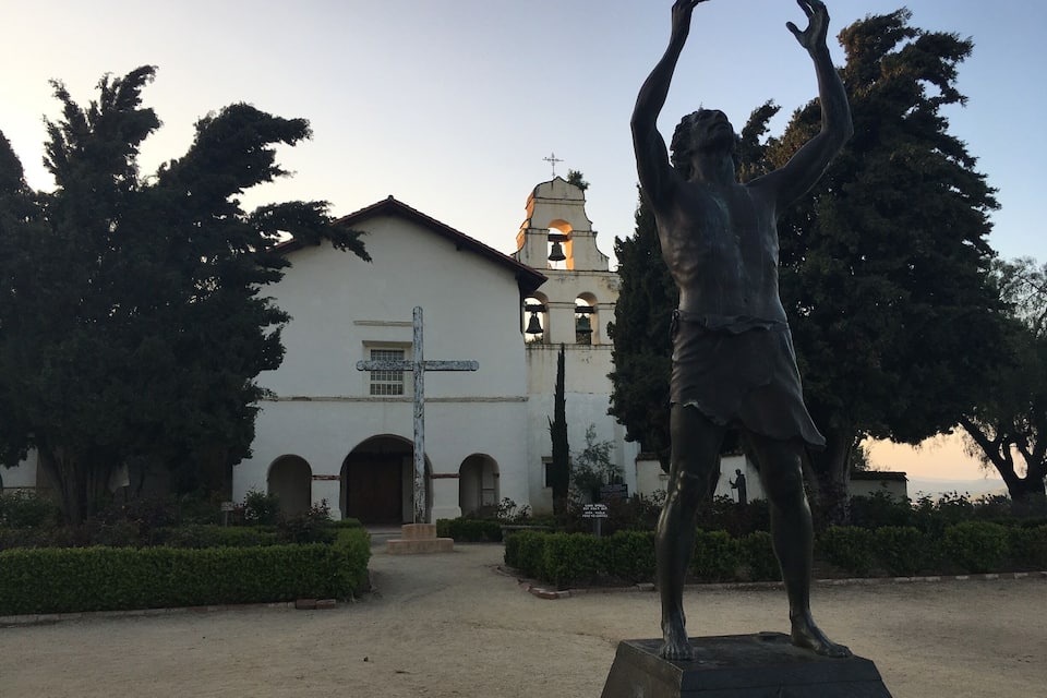 A statue of a man with raised arms in front of a white church with a bell tower.