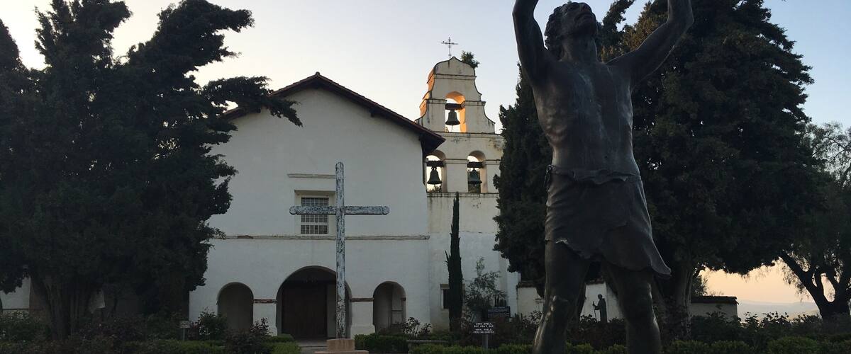 A statue of a man with raised arms in front of a white church with a bell tower.