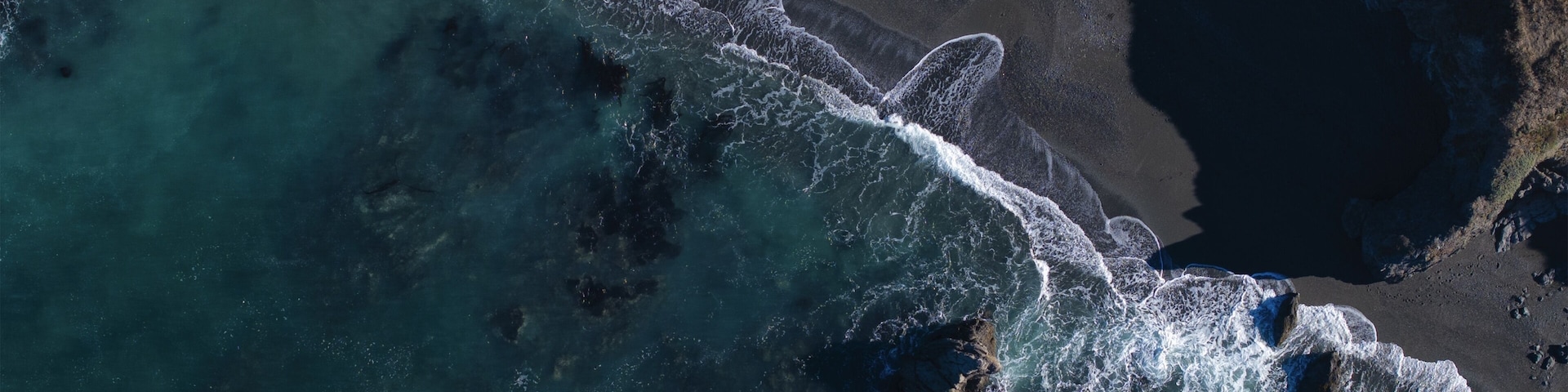 Uma área costeira com ondas quebrando em uma praia arenosa e afloramentos rochosos.