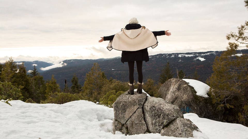 A person standing on a rock with arms outstretched, overlooking a snowy mountainous landscape.
