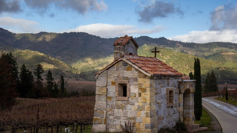 A small stone church with a cross on its roof, surrounded by vineyards and hills.