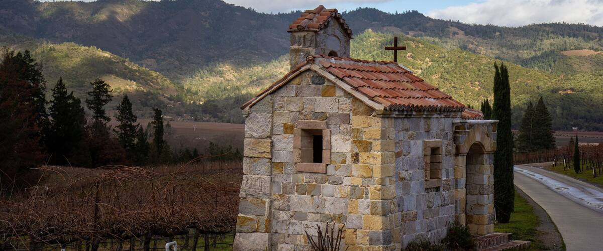 A small stone church with a cross on its roof, surrounded by vineyards and hills.