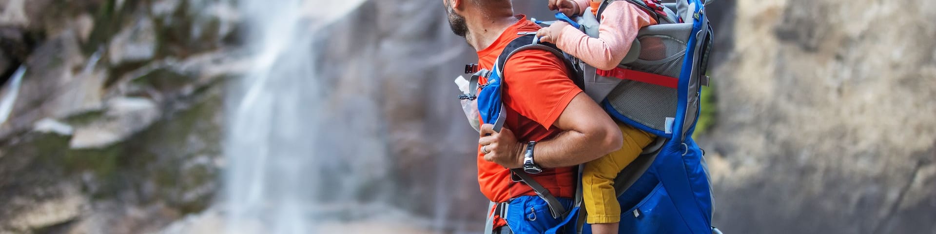 A person carrying a child on their back with a backpack, standing in front of a waterfall.