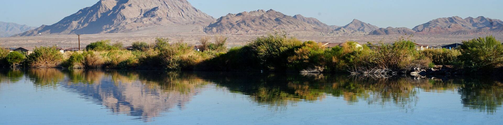 A calm lake with reflections of a mountain range and sparse vegetation in the foreground.