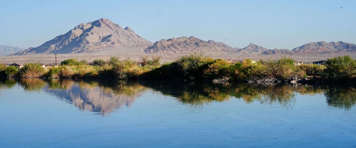 A calm lake with reflections of a mountain range and sparse vegetation in the foreground.