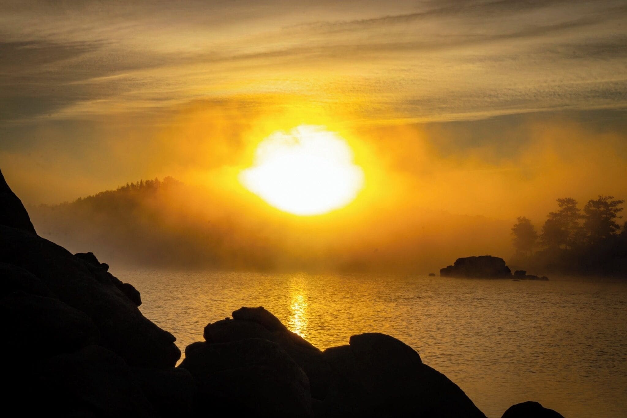 A sunset over a calm body of water with rocky shores and a forested hill in the background.