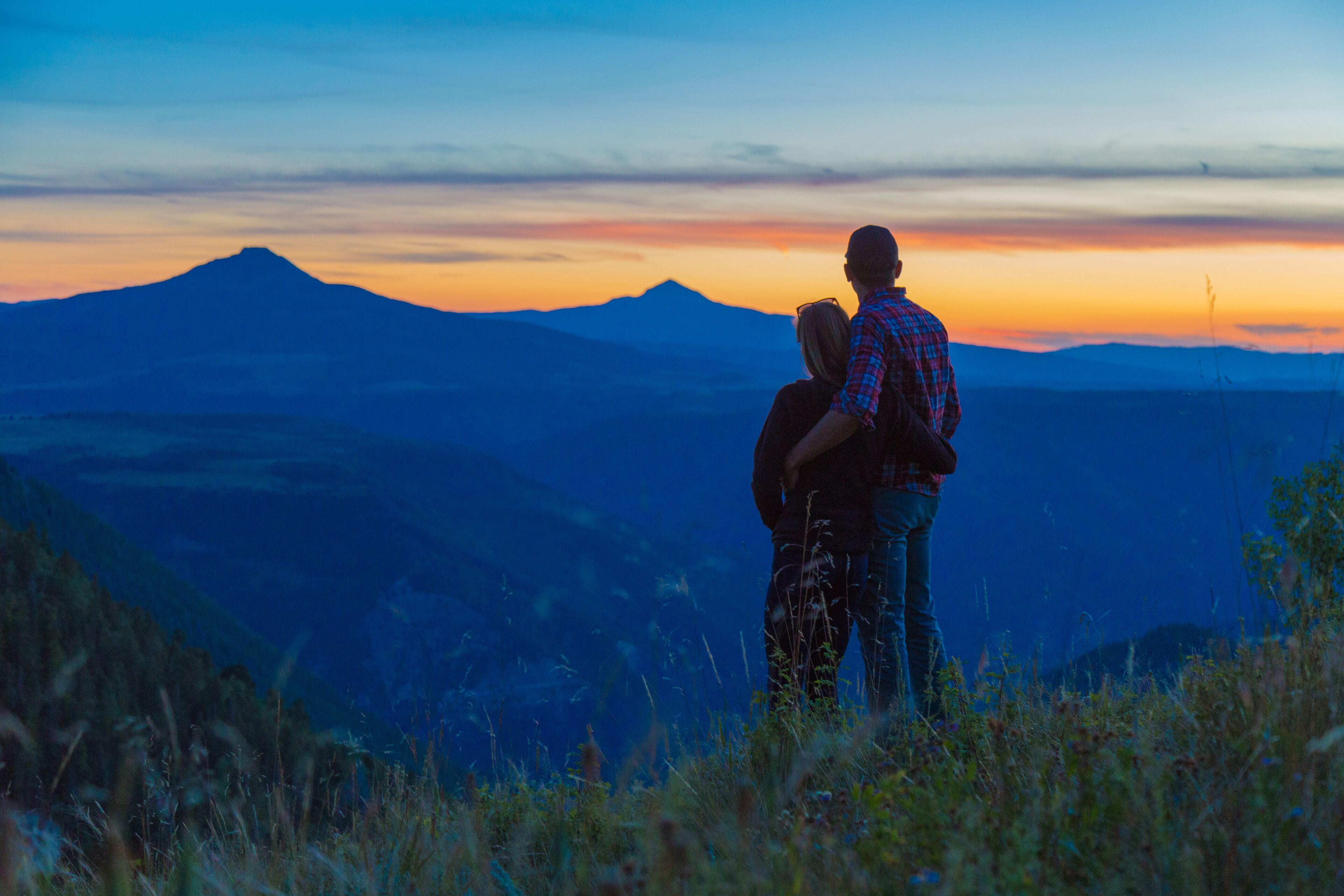 A couple standing on a hill overlooking a mountain range during sunset.