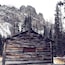 A rustic log cabin with a small window, surrounded by a forest and snow-covered mountains.