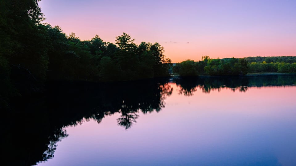A calm lake at sunset with reflections of trees and a clear sky.
