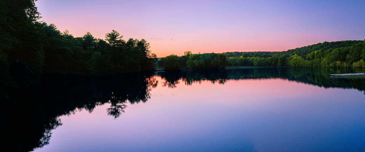 A calm lake at sunset with reflections of trees and a clear sky.