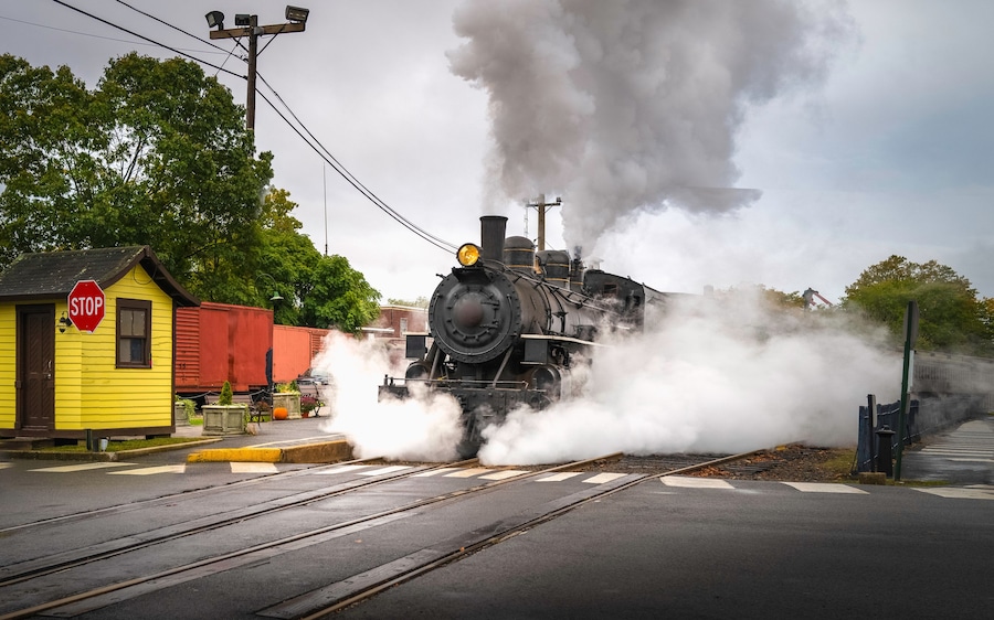 Retro-style historical steam train in motion on the train track in Essex, Connecticut. White steam clouds encircling and rising over the train.
