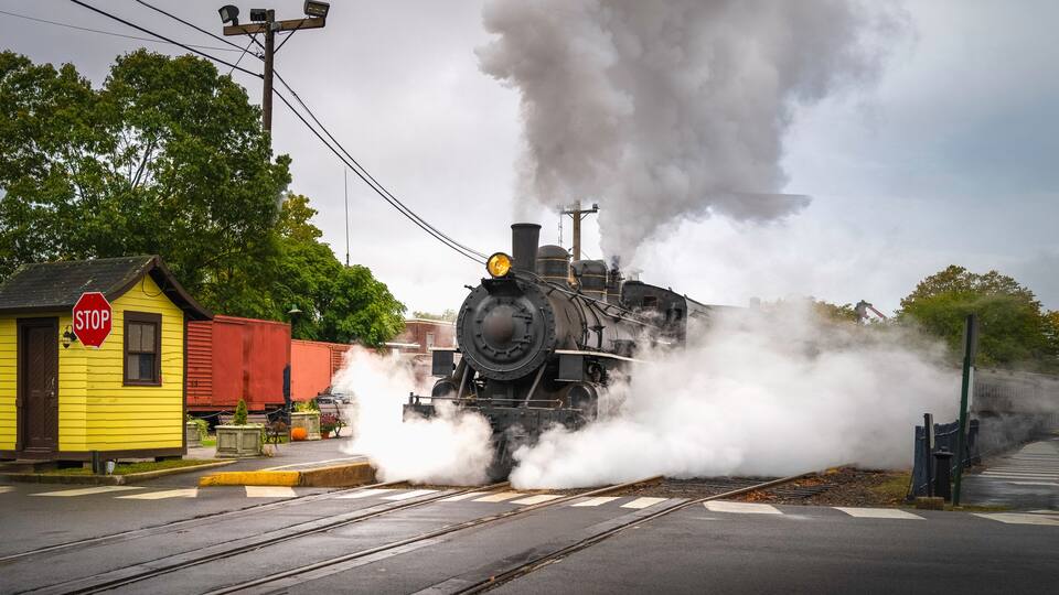 A steam locomotive emitting smoke, a yellow building with a stop sign, and a red train car.