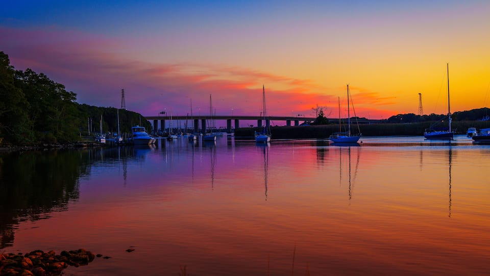 A sunset over a calm harbor with boats and a bridge in the background.
