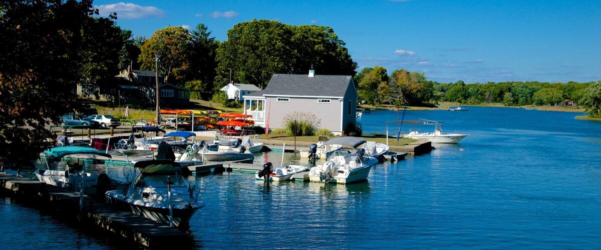 A marina with boats docked, a small white house, and a forested shoreline.