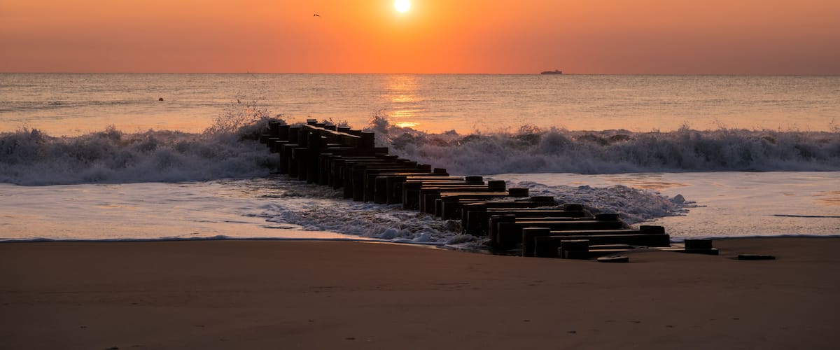 A sunset over the ocean with waves crashing onto a wooden breakwater.