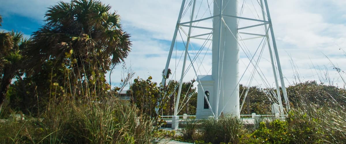 A tall white lighthouse with a metal framework, surrounded by palm trees and greenery.