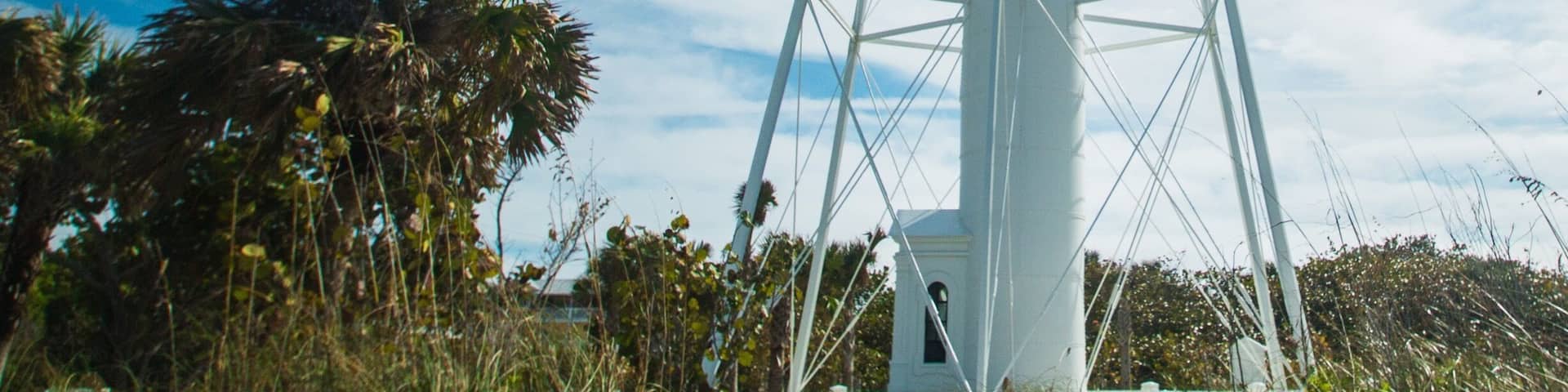 A tall white lighthouse with a metal framework, surrounded by palm trees and greenery.