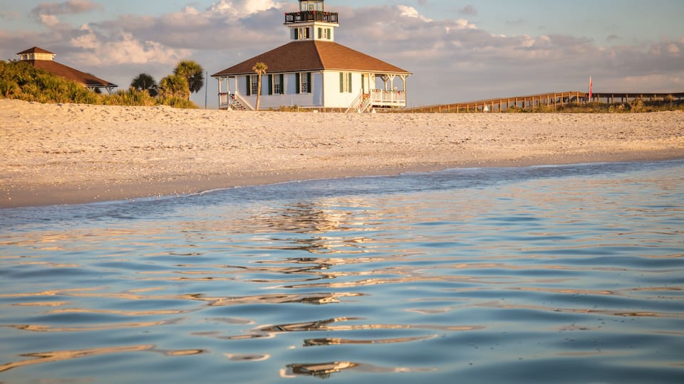 A beach with a white lighthouse, clear blue water, and a wooden pier.