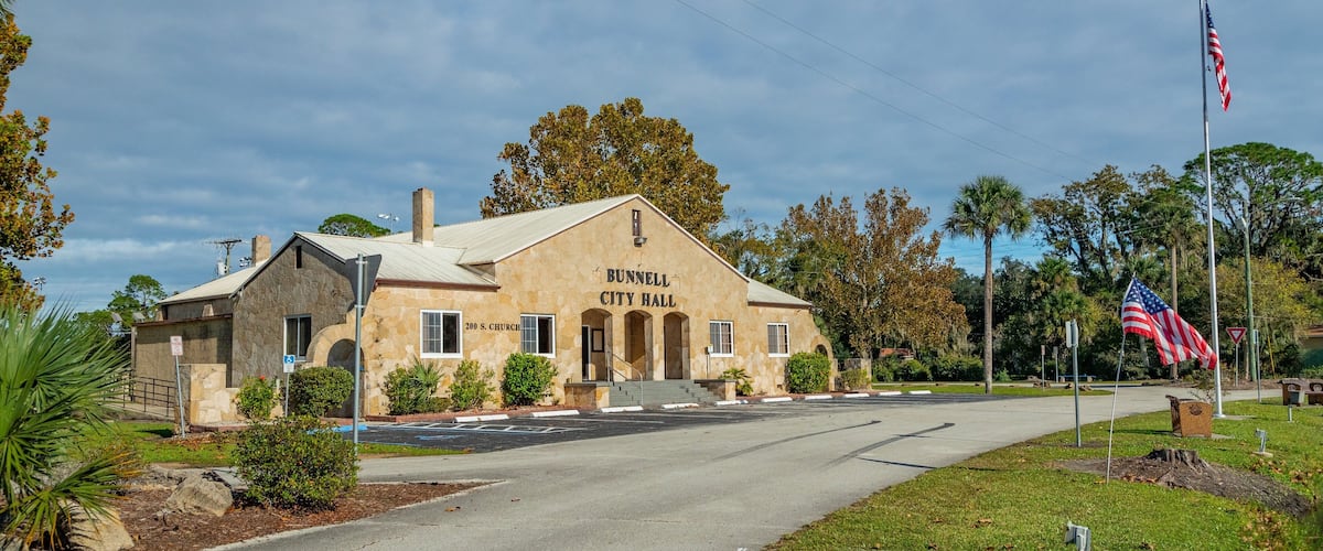 Bunnell City Hall with a stone facade, a flagpole with an American flag, and a street in front.