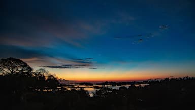 A twilight sky with clouds, a body of water, and silhouettes of trees.