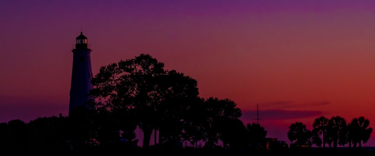 A lighthouse silhouette against a purple sky, surrounded by trees.