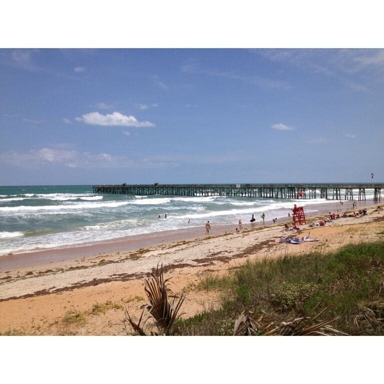 A beach with a pier extending into the ocean, sandy shore, and people enjoying the beach.