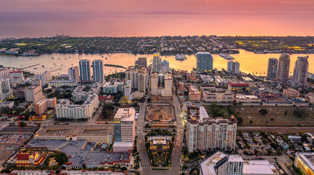 A coastal cityscape at sunset with high-rise buildings, a marina, and a river.