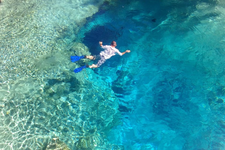 Eine Person schwimmt in klarem, blauem Wasser, wobei Unterwasserfelsen und Korallenformationen sichtbar sind.