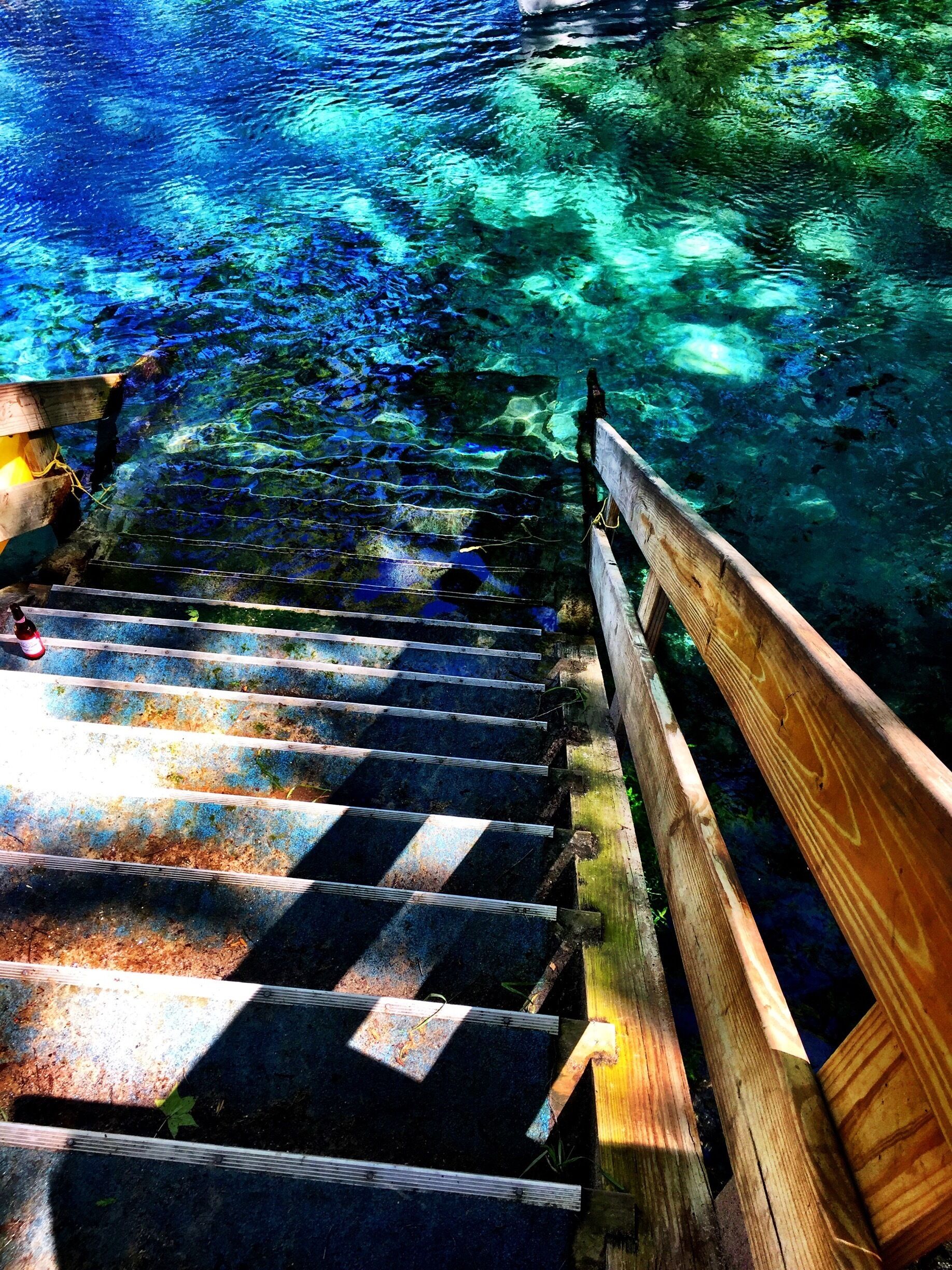 A wooden staircase leading into a clear blue body of water surrounded by greenery.